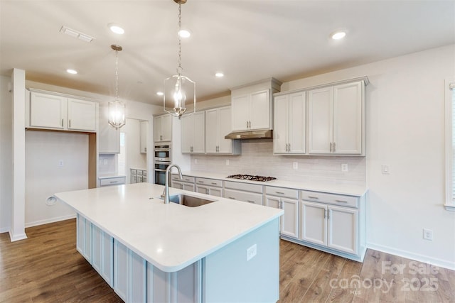 kitchen featuring a sink, a center island with sink, visible vents, baseboards, and under cabinet range hood