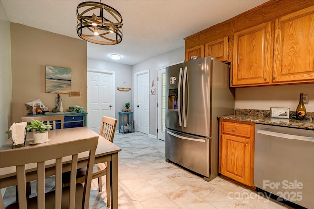 kitchen featuring brown cabinetry, stainless steel appliances, dark stone countertops, a chandelier, and hanging light fixtures