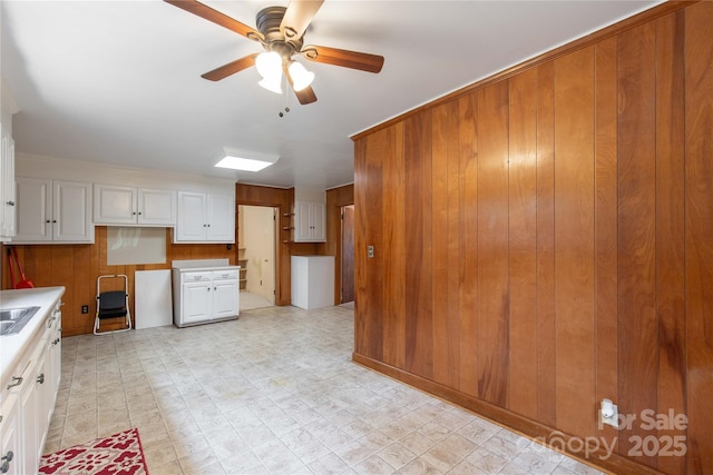 kitchen featuring wooden walls, a ceiling fan, white cabinets, light countertops, and a sink