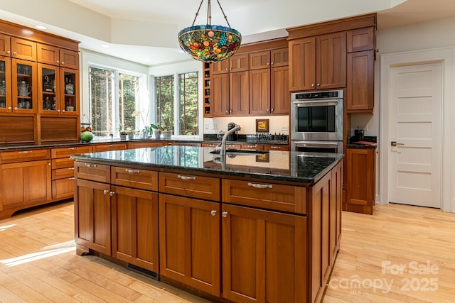kitchen with brown cabinets, double oven, light wood-style flooring, and a kitchen island with sink