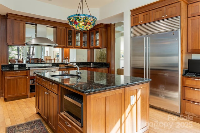 kitchen with a sink, dark stone counters, wall chimney exhaust hood, stainless steel appliances, and light wood-style floors
