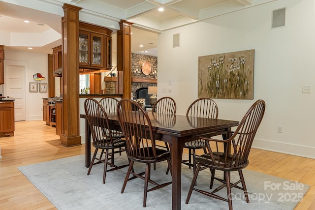 dining space featuring a fireplace, decorative columns, light wood-type flooring, baseboards, and beamed ceiling