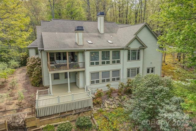 back of house featuring a balcony, a chimney, a forest view, and roof with shingles
