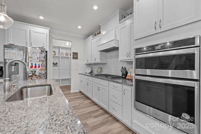 kitchen with a sink, premium range hood, stainless steel appliances, white cabinets, and light stone countertops