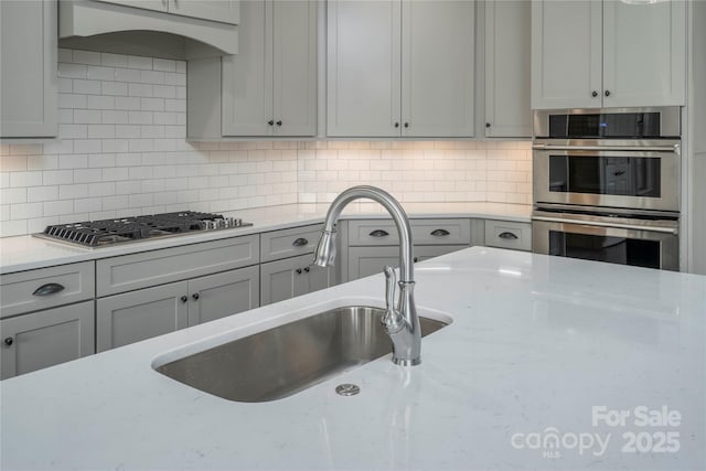 kitchen with a sink, tasteful backsplash, gray cabinets, light stone countertops, and appliances with stainless steel finishes