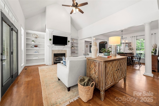 living area featuring high vaulted ceiling, wood finished floors, a tile fireplace, ceiling fan, and decorative columns