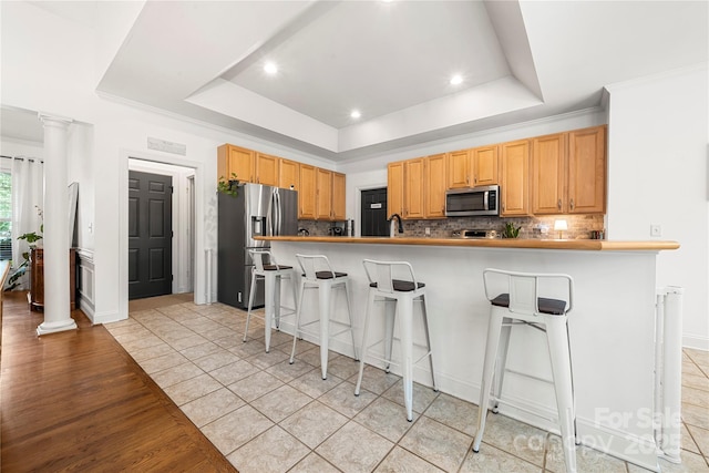 kitchen with stainless steel appliances, decorative columns, a raised ceiling, a breakfast bar area, and decorative backsplash