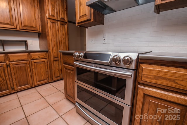 kitchen featuring double oven range, range hood, brown cabinetry, tasteful backsplash, and dark countertops