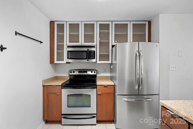 kitchen featuring appliances with stainless steel finishes, brown cabinetry, glass insert cabinets, and light stone countertops