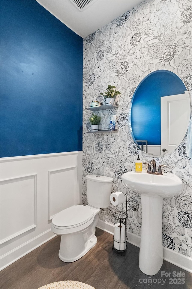 bathroom with wallpapered walls, wainscoting, wood finished floors, toilet, and baseboards