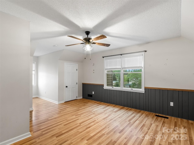 empty room featuring a wainscoted wall, light wood-type flooring, visible vents, ceiling fan, and vaulted ceiling