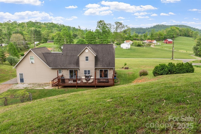 back of property featuring a wooded view, a wooden deck, a shingled roof, and a lawn