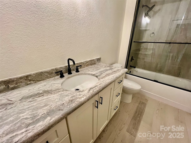 full bath with a textured wall, wood finished floors, vanity, toilet, and enclosed tub / shower combo