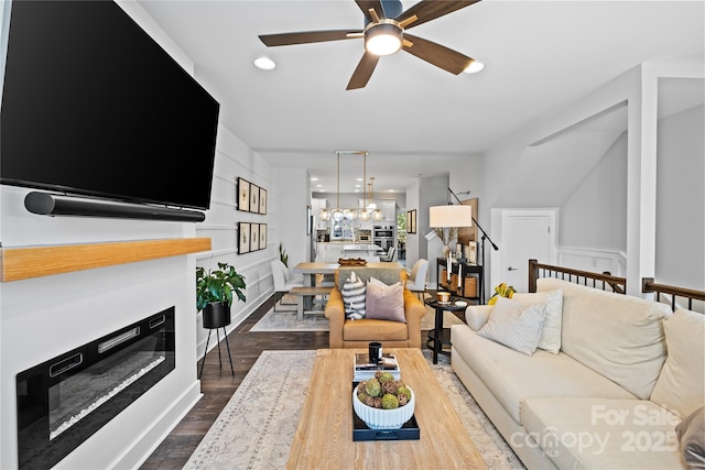 living room with dark wood-style floors, a ceiling fan, a glass covered fireplace, a decorative wall, and a chandelier