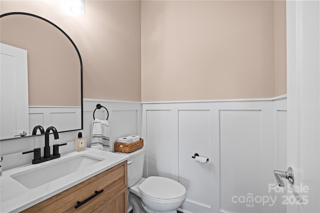 half bath featuring a decorative wall, a wainscoted wall, toilet, and vanity