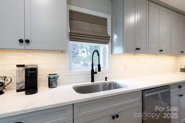 kitchen with a sink, gray cabinets, stainless steel dishwasher, light stone countertops, and backsplash