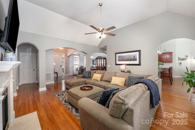 living room with ceiling fan, arched walkways, light wood-style flooring, baseboards, and high vaulted ceiling