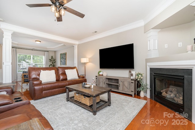 living area with decorative columns, ceiling fan, light wood-type flooring, crown molding, and a glass covered fireplace