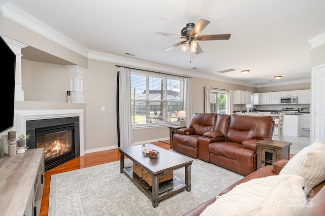 living area with ornamental molding, a ceiling fan, light wood-style flooring, a glass covered fireplace, and baseboards