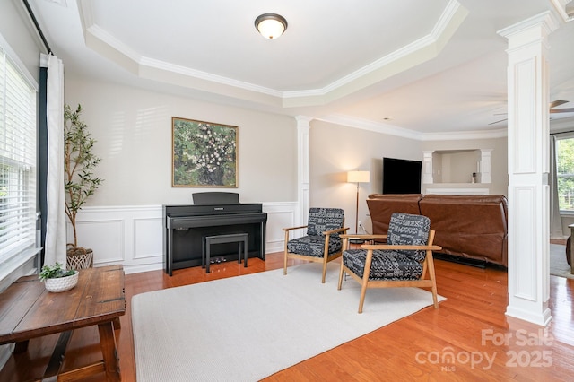 sitting room featuring ornate columns, a raised ceiling, wood finished floors, ornamental molding, and wainscoting