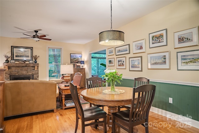 dining space featuring healthy amount of natural light, light wood-style flooring, a ceiling fan, a fireplace, and wainscoting