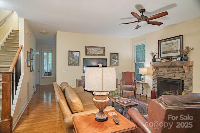 living room with stairs, light wood-style floors, a ceiling fan, a stone fireplace, and baseboards