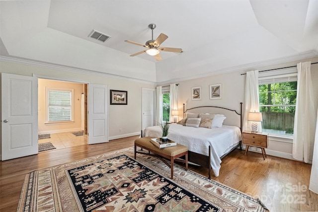 bedroom featuring light wood-type flooring, a raised ceiling, multiple windows, baseboards, and crown molding