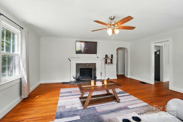 living room featuring arched walkways, light wood-style floors, a fireplace, a ceiling fan, and ornamental molding