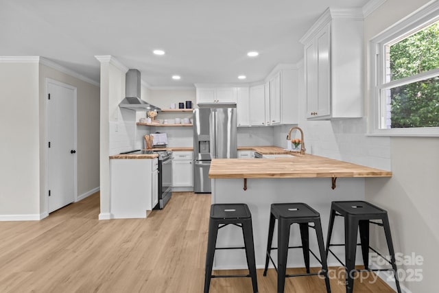 kitchen with wood counters, appliances with stainless steel finishes, a sink, wall chimney exhaust hood, and a peninsula