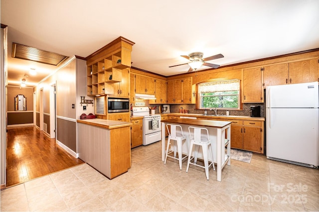 kitchen with white appliances, under cabinet range hood, open shelves, brown cabinetry, and crown molding