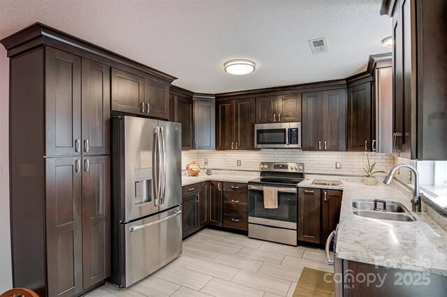 kitchen featuring stainless steel appliances, a sink, dark brown cabinets, tasteful backsplash, and light stone counters