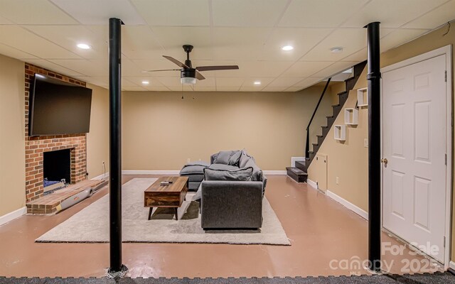 living room with finished concrete flooring, baseboards, a brick fireplace, ceiling fan, and a drop ceiling