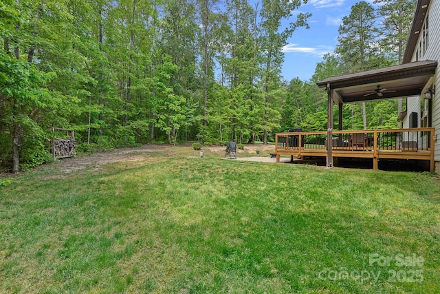 view of green lawn featuring ceiling fan, a wooden deck, and a forest view