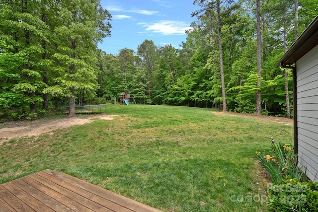 view of grassy yard with a trampoline, a playground, a view of trees, and a deck