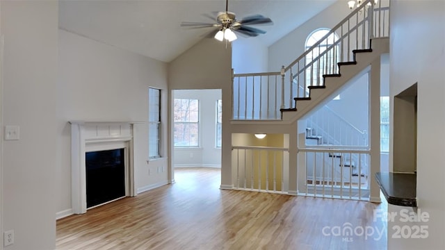 unfurnished living room with a ceiling fan, wood finished floors, high vaulted ceiling, stairway, and baseboards