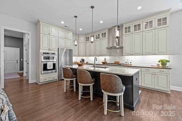 kitchen featuring stainless steel appliances, wall chimney exhaust hood, a sink, dark wood-style floors, and backsplash