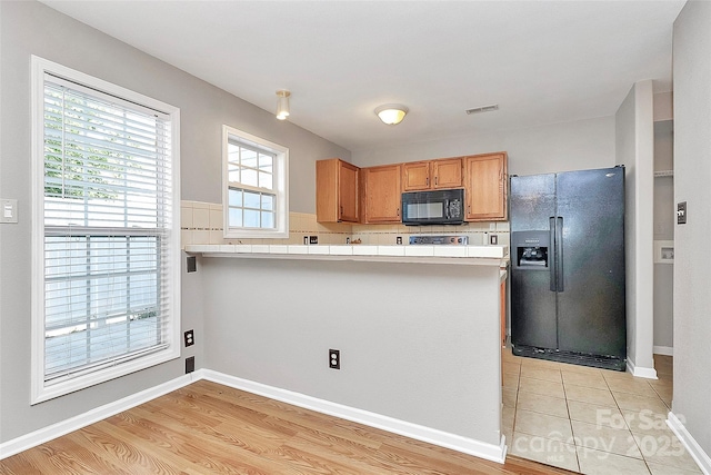 kitchen with black appliances, a peninsula, tile countertops, light wood-type flooring, and baseboards