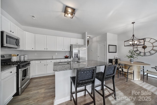 kitchen featuring appliances with stainless steel finishes, a sink, wood finished floors, dark stone countertops, and an island with sink