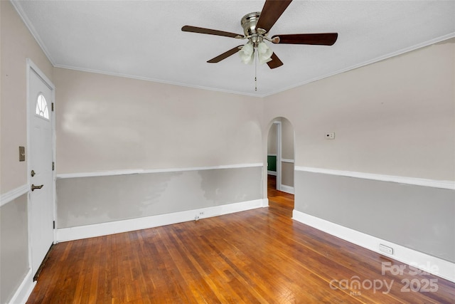 unfurnished room featuring ceiling fan, arched walkways, wood-type flooring, ornamental molding, and baseboards