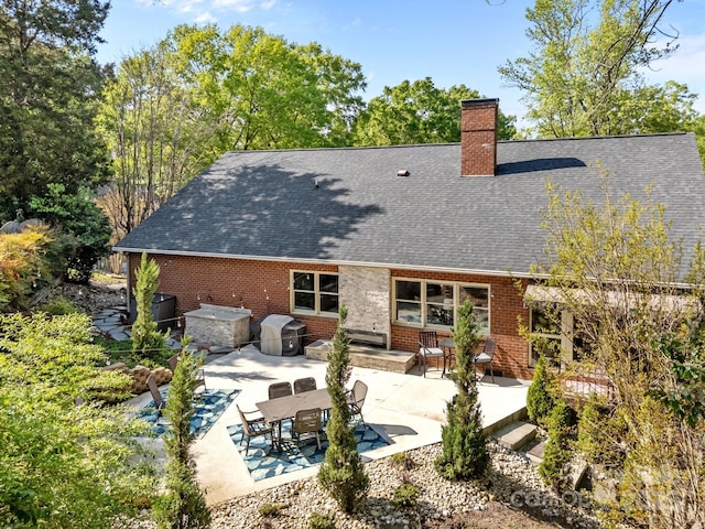 back of property featuring brick siding, a patio, roof with shingles, and a chimney