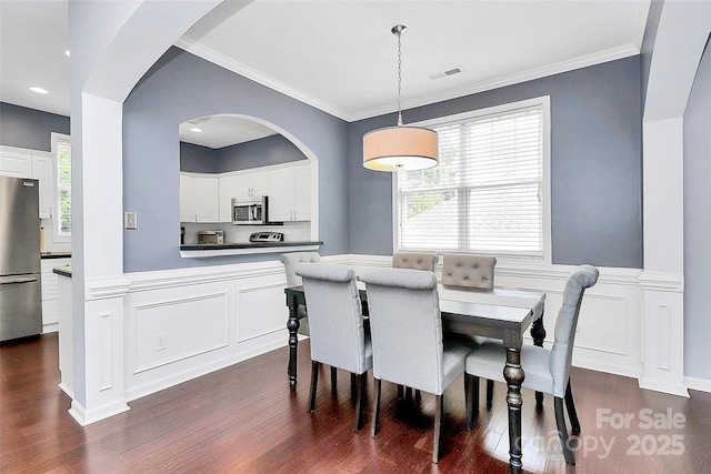 dining area with dark wood finished floors, a wainscoted wall, a decorative wall, crown molding, and arched walkways