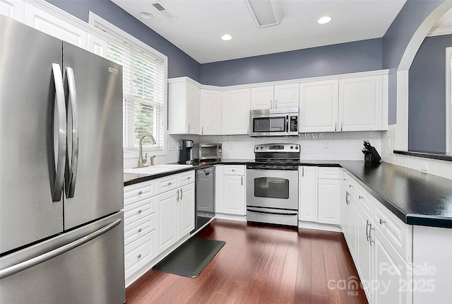 kitchen featuring stainless steel appliances, a sink, dark countertops, arched walkways, and dark wood-type flooring