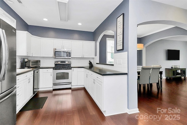 kitchen with stainless steel appliances, arched walkways, dark wood-style flooring, dark countertops, and tasteful backsplash