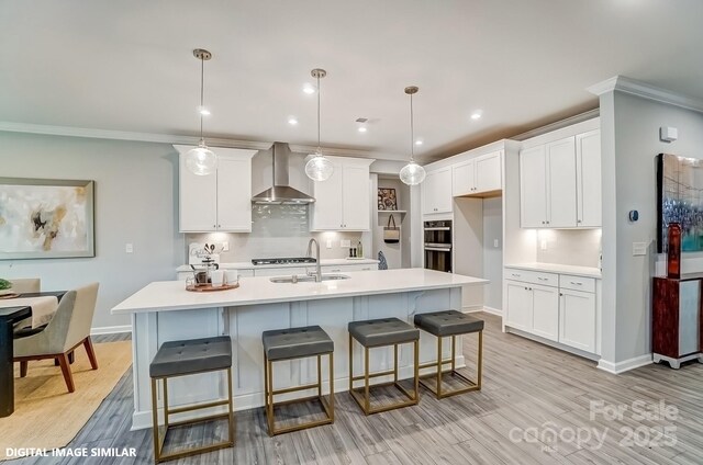 kitchen featuring wall chimney range hood, a sink, light countertops, white cabinets, and crown molding