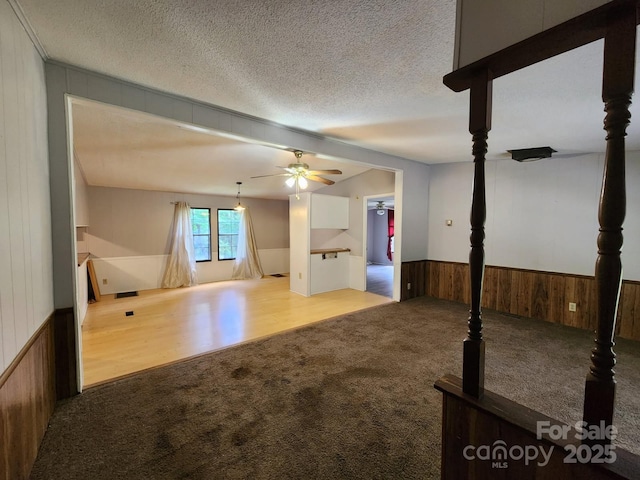 unfurnished living room with carpet flooring, wainscoting, a ceiling fan, wooden walls, and a textured ceiling