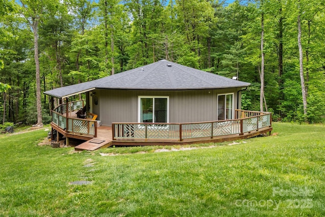 rear view of property featuring roof with shingles, a wooden deck, a view of trees, and a lawn