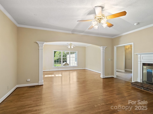 unfurnished living room featuring a ceiling fan, wood finished floors, ornate columns, a brick fireplace, and ornamental molding