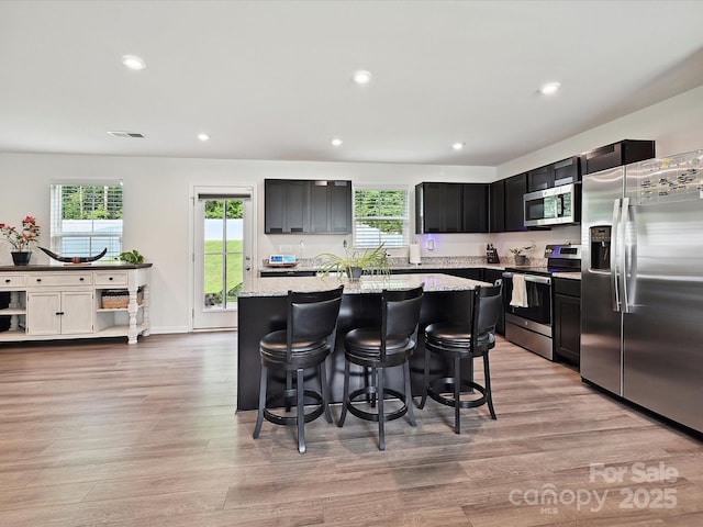 kitchen featuring appliances with stainless steel finishes, a center island, dark cabinetry, a kitchen bar, and light stone countertops