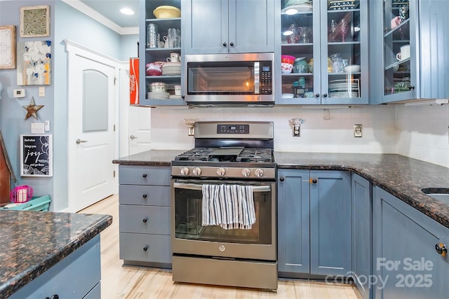 kitchen with appliances with stainless steel finishes, backsplash, dark stone counters, glass insert cabinets, and blue cabinetry