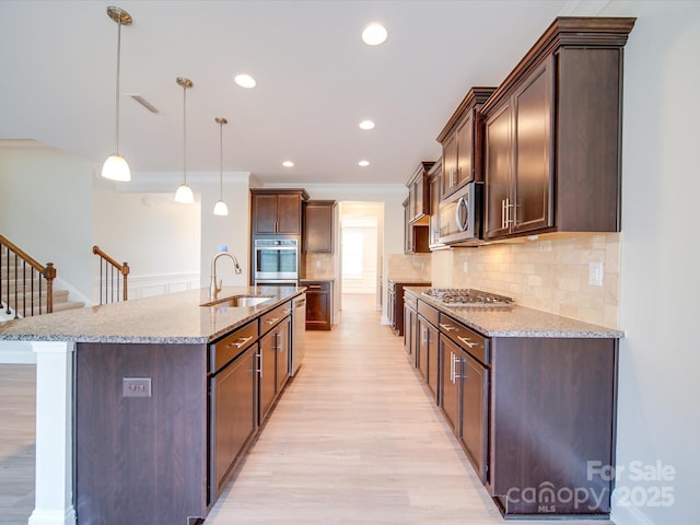 kitchen featuring a sink, stainless steel appliances, light wood-style flooring, a center island with sink, and light stone counters